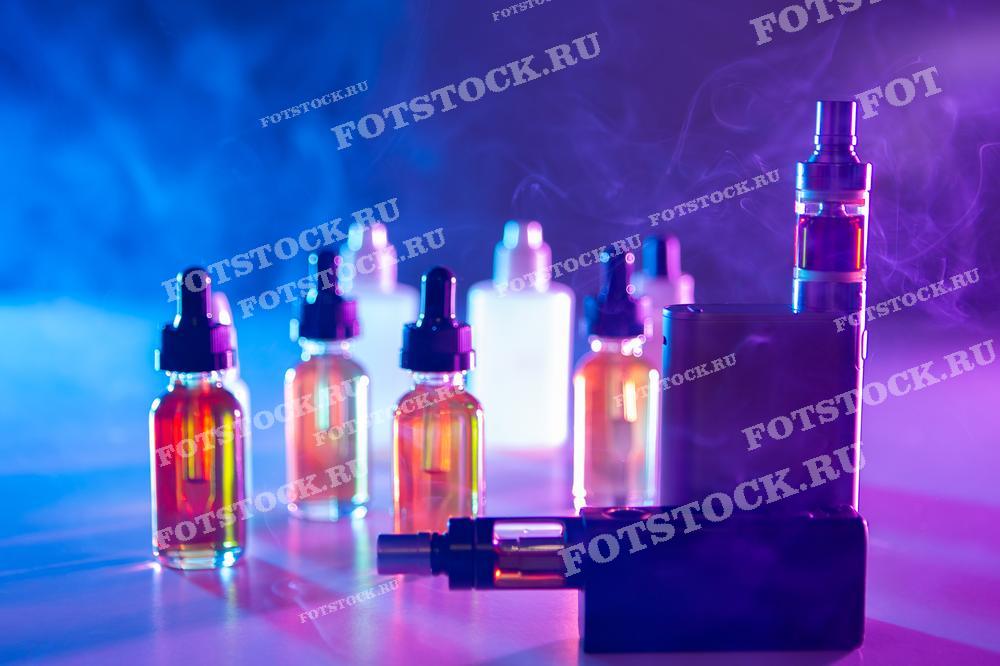 На фотографии изображены несколько вейп-устройств и флаконов с жидкостью для вейпинга, расположенных на поверхности на фоне густого цветного дыма. Снимок подчеркивает современный стиль и разнообразие оборудования для вейпинга.